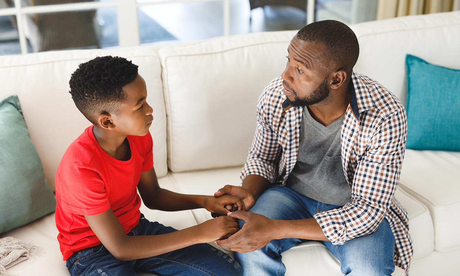 Collaborative Problem Solving (Serious african american father and son sitting on couch in living room talking and holding hands)