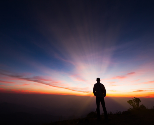 Silhouette of a man standing on the cliff looking at sunrise background, hope and following a dream concept